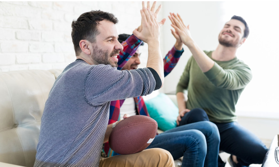 Three guys sitting on a couch celebrating by giving high-fives, while one is holding a football.
